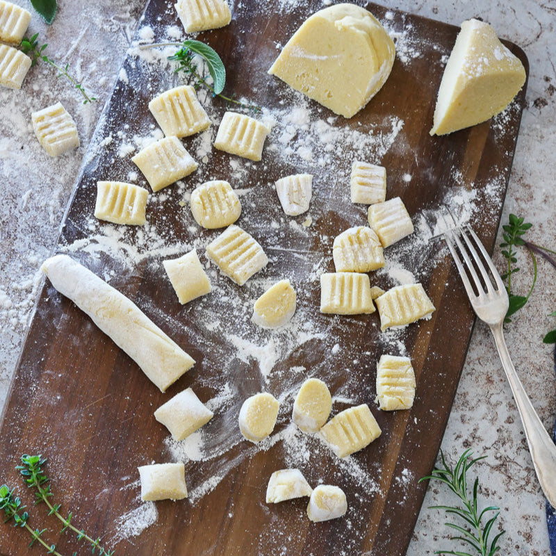 Die ungekochten Ruut glutenfrei Gnocchi liegen auf einem bemehlten Holzbrett, auf dem eine Gabel und frische Kräuter verstreut sind.