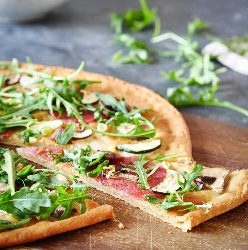 Eine glutenfreie Ruut-Pizza mit dünner Kruste, belegt mit Rucola, Zucchini und Salami, serviert auf einem Holzbrett.