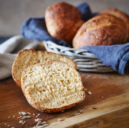 Ein Ruut Brötchen, ohne Zusatzstoffe, ist auf ein Holzbrett aufgeschnitten, während ganze Brötchen im Korb dahinter liegen.